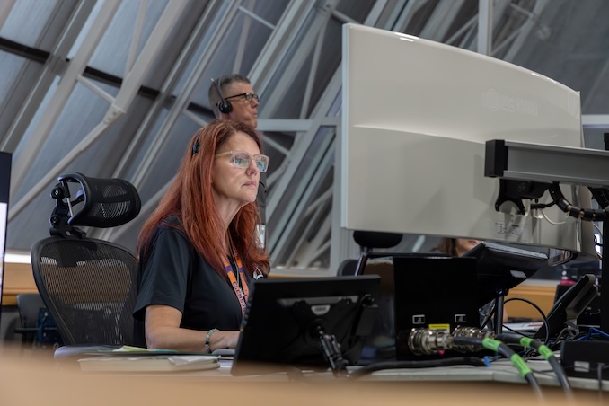 Two people wearing headsets work together at computer stations inside a control room setting, with one person seated at a keyboard and the other person standing nearby.